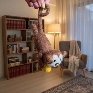 Può includere: Una scimmia fatta a mano in feltro marrone con viso bianco, occhi neri e una palla gialla. La scimmia è appesa a testa in giù. Lo sfondo include una libreria, una lampada e una sedia.