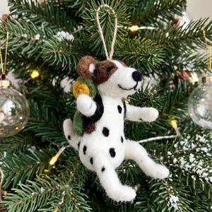 May include: A handmade felt dog Christmas ornament. The dog is white with black spots, brown ears, and a black nose. It has a green and yellow backpack and is hanging from a natural twine string. The ornament is set against a backdrop of a Christmas tree.