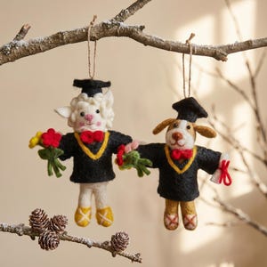 May include: Two felt graduation ornaments: a sheep and a dog. Both wear black graduation gowns with gold trim, red bow ties, and mortarboards. The sheep holds a bouquet of flowers, while the dog holds a diploma. They hang from a snowy branch.