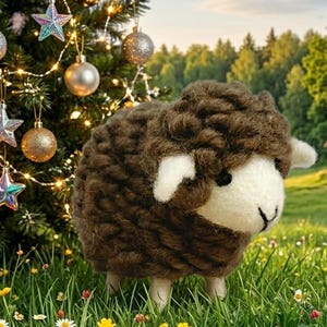 May include: A small, handcrafted sheep figurine made of textured brown yarn with a smooth white felt face, black bead eyes, and a stitched nose, standing in a grassy field near a decorated Christmas tree with ornaments.