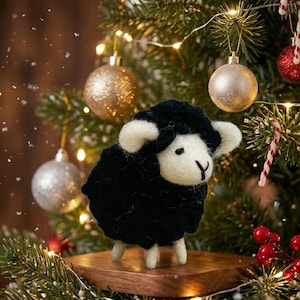 May include: A needle-felted sheep figurine with a black wool body and white wool face, ears, and legs. The sheep stands on a small wooden platform, positioned in front of a green pine tree decorated with gold, silver, and red ornaments.
