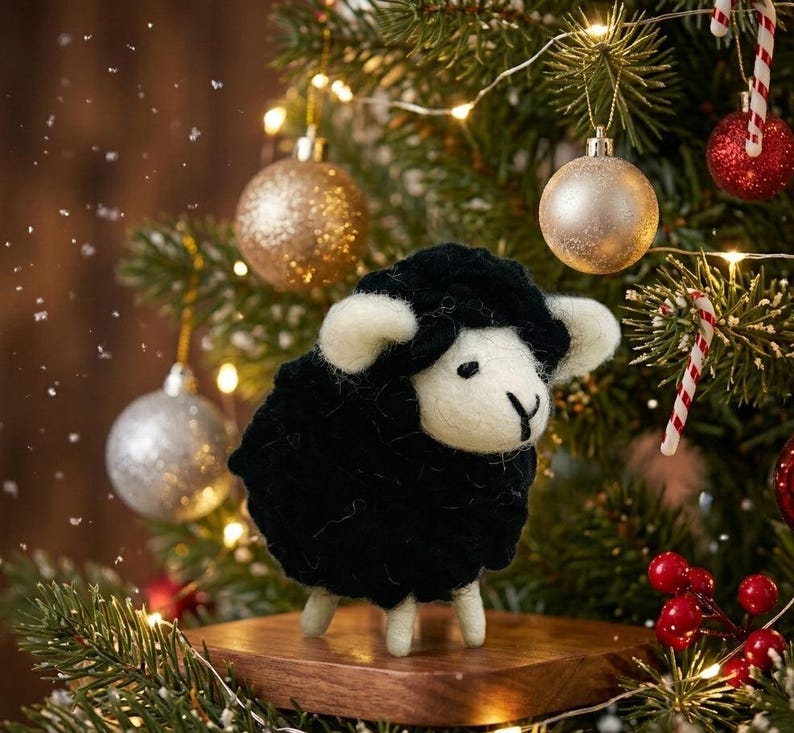 May include: A needle-felted sheep figurine with a black wool body and white wool face, ears, and legs. The sheep stands on a small wooden platform, positioned in front of a green pine tree decorated with gold, silver, and red ornaments.