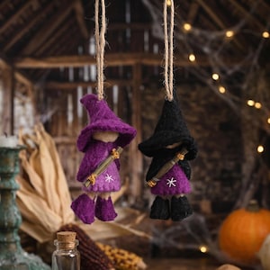 Op de afbeelding: Twee vilten heksenornamenten die aan touw hangen. De ene heks is paars met een bijpassende hoed en laarzen, de andere zwart met een bijpassende hoed en laarzen. Beide heksen hebben een ster op hun jurk en een bezem.
