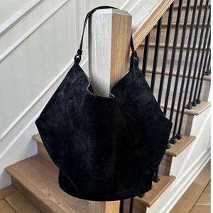 May include: A black suede tote bag with a single shoulder strap. The bag has a distinctive, angular shape and a soft, velvety texture. The bag is displayed on a staircase with wooden steps and a black metal railing.