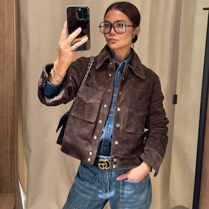 May include: A brown suede jacket with four front pockets and button closures. The jacket is worn over a denim shirt and paired with blue jeans. A gold-buckled belt and a black shoulder bag complete the look. The person is wearing glasses and holding a phone.
