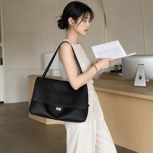 以下が含まれることがあります： 長方形のシルエット、折りたたみ式のフラップ、金属製のターンロック式開閉部を備えた大きな黒いレザーショルダーバッグ。2本の長く細いショルダーストラップと質感のある表面が特徴で、構造化された外観を提供します。
