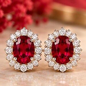 May include: A pair of stud earrings featuring large oval-cut deep red center stones surrounded by a halo of small round white stones set in a gold-toned metal frame. The earrings have a classic prong setting and a polished finish.