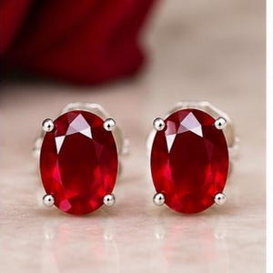 Puede incluir: Un par de pendientes de botón con gemas de color rojo intenso de corte ovalado engastadas en metal plateado pulido. Cada piedra está asegurada por cuatro pequeñas garras en los lados, mostrando una superficie facetada que refleja la luz.