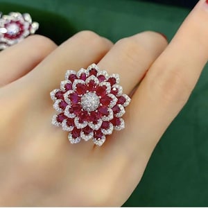 Puede incluir: Un anillo con forma de flor que presenta múltiples capas de rubíes de color rojo intenso en talla pera dispuestos en un patrón de pétalos. El centro es un grupo de pequeños diamantes blancos redondos y cada rubí está delineado con diamantes.