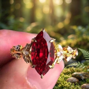 Pode incluir: Um anel de ouro com uma grande pedra preciosa vermelho-escura em lapidação marquise como pedra central, ladeada por pedras transparentes menores em lapidação marquise dispostas num padrão de folhas ao longo do aro em metal dourado.