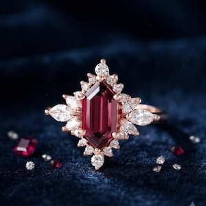 Pode incluir: Um anel de ouro rosa com uma pedra preciosa central de cor vermelho-escura em formato hexagonal alongado, rodeada por um grupo de pequenos diamantes brancos em lapidação marquise e brilhante. O anel é acompanhado por pedras soltas ao redor.
