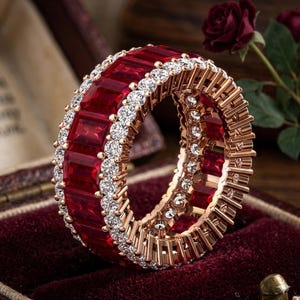 May include: A rose gold eternity band featuring a central row of rectangular, deep red gemstones flanked by two rows of small, round, clear, sparkling stones. The ring has a continuous, uniform design with a metallic finish and vibrant red colour.