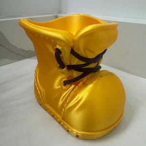 Op de afbeelding: Een felgele, 3D-geprinte laarzenvormige plantenbak. De laars heeft een ronde neus, een veter aan de voorkant met donkerbruine veters en een textuurzool. De opening van de plantenbak bevindt zich aan de bovenkant en het materiaal heeft een satijnachtige glans.