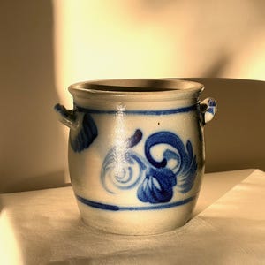 Puede incluir: Una vasija de gres cilíndrica con una base de color gris claro y patrones florales de volutas pintados a mano en azul cobalto. El recipiente incluye dos pequeñas asas laterales y una banda azul horizontal que rodea la parte inferior.