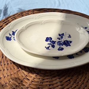 Peut inclure: Deux plats ovales en céramique de couleur crème avec une bordure en relief décorative et un motif floral bleu. Le plus petit plat est centré sur le plus grand, tous deux présentant des grappes de fleurs et des feuilles bleues assorties.