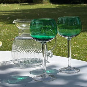 Peut inclure: Deux verres à vin avec des pieds transparents et des bols vert foncé sont placés à côté d'une carafe en verre transparent avec un motif côtelé vertical et un dessin de feuille gravé. La verrerie est disposée sur une surface blanche.