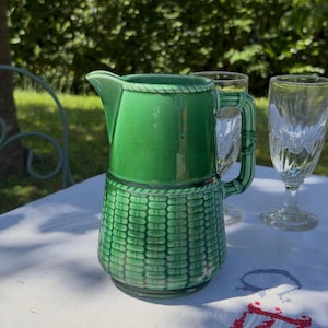 Peut inclure: Une carafe en céramique vert brillant avec une partie supérieure lisse et une base texturée en motif de vannerie tressée. La carafe présente un bord en forme de corde et une anse imitant le bambou, accompagnée de deux verres transparents.