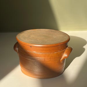 Peut inclure: Un pot en céramique vernissée marron de forme ronde avec un couvercle plat et deux anses latérales. Le récipient présente des détails nervurés horizontaux autour du corps, une surface supérieure lisse et une finition brillante.