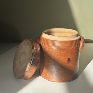 Peut inclure: Un pot cylindrique en grès brun avec son couvercle assorti posé sur le côté. Le récipient présente une finition texturée et tachetée, un bord large et deux petites anses latérales. La surface montre des variations de couleur et une tache.