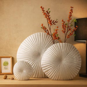 Puede incluir: Tres jarrones circulares texturizados de color blanquecino de diferentes tamaños están dispuestos sobre una superficie de madera. Los jarrones tienen un diseño radial. Ramas de bayas naranjas se colocan en el jarrón más grande. Una imagen enmarcada y objetos decorativos también están en la superficie.