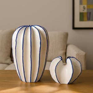 Peut inclure: Deux vases en céramique blanche avec des détails bleus. Le plus grand est cylindrique, le plus petit en forme de cœur. Les deux ont des rayures bleues verticales. Ils sont posés sur une surface en bois.