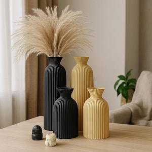 Puede incluir: Colección de jarrones decorativos en negro y amarillo. Los jarrones tienen un diseño acanalado y están colocados sobre una mesa de madera. Un jarrón negro alto contiene hierba de la pampa seca. Dos pequeñas esculturas están frente a los jarrones.