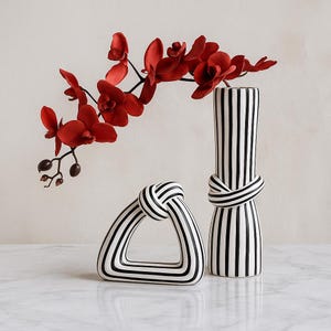 Peut inclure: Un vase rayé noir et blanc avec un motif de nœud, à côté d'un objet décoratif triangulaire avec un motif assorti. Des fleurs d'orchidées rouges sont disposées dans le vase. Les objets sont sur une surface blanche.