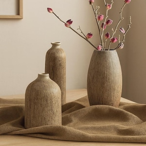 Peut inclure: Trois vases bruns texturés de hauteurs différentes sont présentés sur une surface en bois, avec un tissu brun drapé devant. Le vase le plus grand contient des branches avec des bourgeons roses. Les vases ont une esthétique neutre et terreuse.