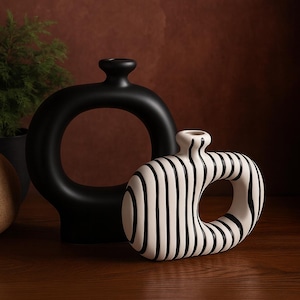 Peut inclure: Deux vases en céramique. L'un est noir et en forme d'anneau, avec une ouverture circulaire au centre. L'autre est blanc avec des rayures verticales noires et une ouverture ovale. Les deux vases ont un design moderne et minimaliste.