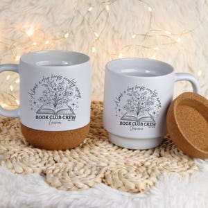 Könnte beinhalten: Zwei weiße Keramikbecher mit Korkböden, mit einem Blumen- und Buchdesign und dem Text "A book a day keeps reality away" und "Book Club Crew". Ein Becher hat einen Deckel. Die Becher stehen auf einer gewebten Matte.