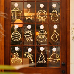 Puede incluir: Colección de adornos navideños iluminados para ventanas, que incluyen un regalo, una estrella, una corona, un muñeco de nieve, una bola, un reno, Papá Noel, un calcetín, acebo, un bastón de caramelo, un árbol de Navidad y un gorro. Luces blancas cálidas.