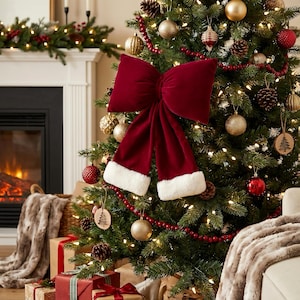 May include: A Christmas tree decorated with gold and red baubles, pinecones, and a red beaded garland. A large, plush, burgundy bow with white trim is the focal point. Presents are at the base of the tree, creating a festive holiday scene.