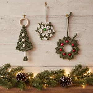 Puede incluir: Tres adornos navideños de macramé hechos a mano: un árbol verde con cuentas de madera, una corona blanca y verde y una corona verde con cuentas rojas. Los adornos cuelgan de una pared de madera blanca. Ramas de pino y piñas en la parte inferior.