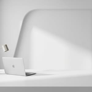 Può includere: Un'area di lavoro bianca minimalista con un laptop con il logo Apple, una lampada da scrivania e un piccolo vaso con una pianta. La scena è immersa nella luce naturale, creando un'estetica pulita e moderna. La scrivania è una semplice superficie bianca.