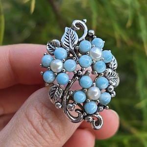May include: A silver-toned ring featuring a floral design with light blue round stones and white pearls. The metal setting is shaped like leaves and vines, creating a cluster of flowers. The ring has a polished finish and intricate metalwork details.