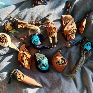 Puede incluir: Una colección de colgantes hechos a mano de cuero y tela en varias formas y colores. Los colgantes presentan diseños intrincados, incluyendo caras y formas abstractas, con acentos azules, marrones y beige. Algunos colgantes tienen detalles de cuentas o trenzados.