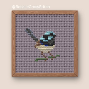 May include: Cross-stitch artwork of a small bird with blue, black, and tan feathers perched on a green branch. The bird is set against a lavender background within a wooden frame. The text "@RosalieCrossStitch" is in the top left corner.