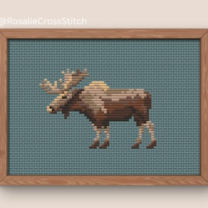 Puede incluir: Obra de arte de punto de cruz de un alce en tonos marrón, beige y blanco, sobre un fondo verde azulado. La pieza enmarcada presenta el texto "@RosalieCrossStitch" en la esquina superior izquierda. El alce se representa de perfil.