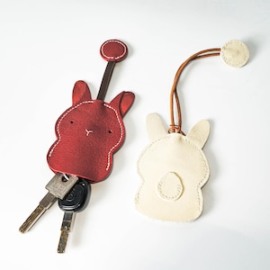 May include: Two leather bunny keychains. One is red with keys attached, the other is off-white. Both have stitched details and a leather strap. The red keychain has a dark brown strap and a circular accent. The off-white keychain has a light brown strap and a circular accent.