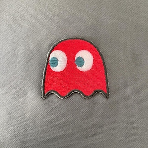 May include: A red embroidered patch in the shape of a ghost, featuring white eyes with teal pupils. The patch has a black border with a wavy bottom edge, set against a gray fabric background. This patch is a fun accessory.