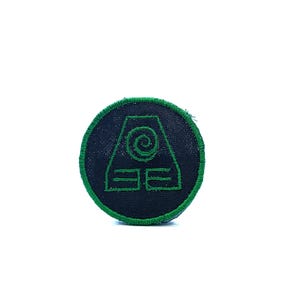 Può includere: Toppa rotonda con sfondo nero e bordo verde. La toppa presenta un simbolo verde con una spirale e due linee orizzontali, probabilmente rappresentante un elemento immaginario. La toppa è probabilmente per abbigliamento o accessori.