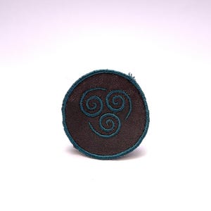 Könnte beinhalten: Ein kreisförmiger Aufnäher mit braunem Zentrum und türkisfarbenem Rand. Der Aufnäher zeigt ein türkisfarbenes Luftnomaden-Symbol, ein stilisiertes Wirbeldesign. Der Aufnäher ist wahrscheinlich für Kleidung oder Accessoires.