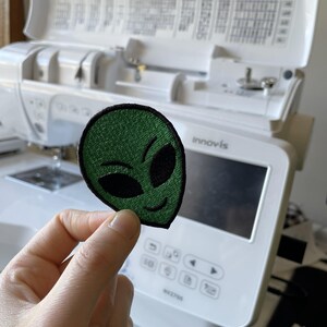 K&ouml;nnte beinhalten: Ein gr&uuml;ner Alien-Aufn&auml;her mit schwarzen Stickdetails. Der Aufn&auml;her zeigt ein l&auml;chelndes Alien-Gesicht. Der Aufn&auml;her wird vor einer wei&szlig;en N&auml;hmaschine gehalten.