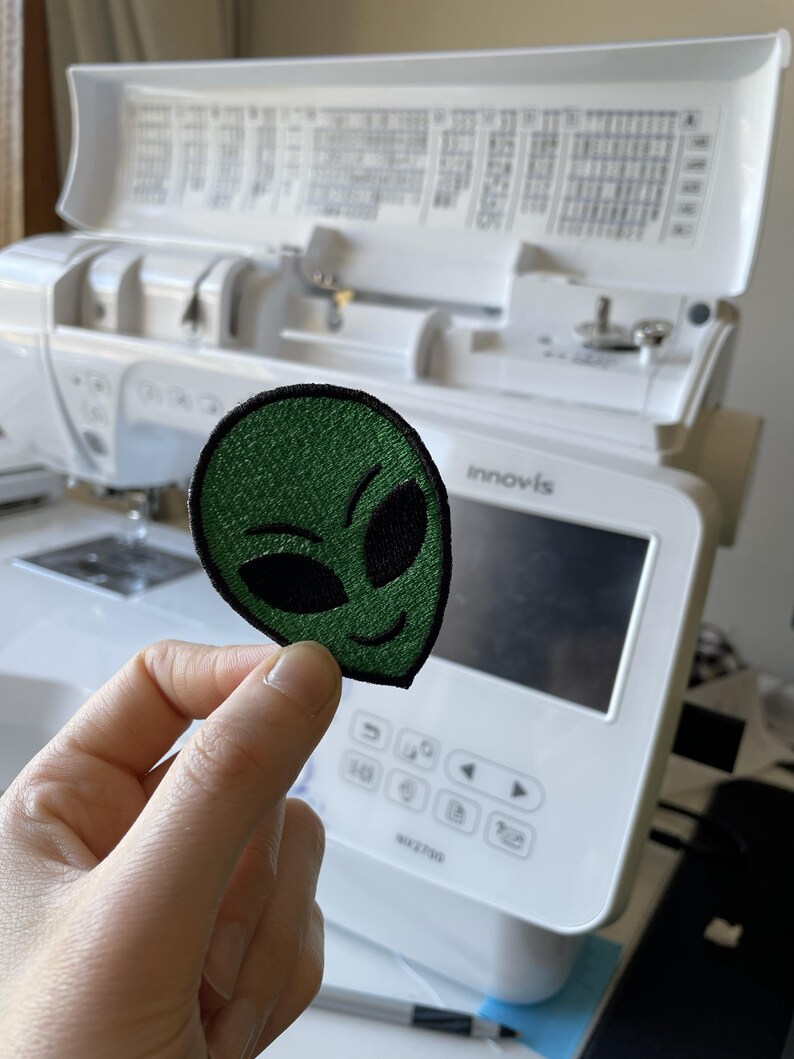 K&ouml;nnte beinhalten: Ein gr&uuml;ner Alien-Aufn&auml;her mit schwarzen Stickdetails. Der Aufn&auml;her zeigt ein l&auml;chelndes Alien-Gesicht. Der Aufn&auml;her wird vor einer wei&szlig;en N&auml;hmaschine gehalten.