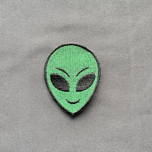Puede incluir: Parche bordado con la cara de un alienígena verde con ojos negros y una boca sonriente. El parche tiene un borde negro y está sobre un fondo de tela gris. El diseño alienígena es un tema popular.