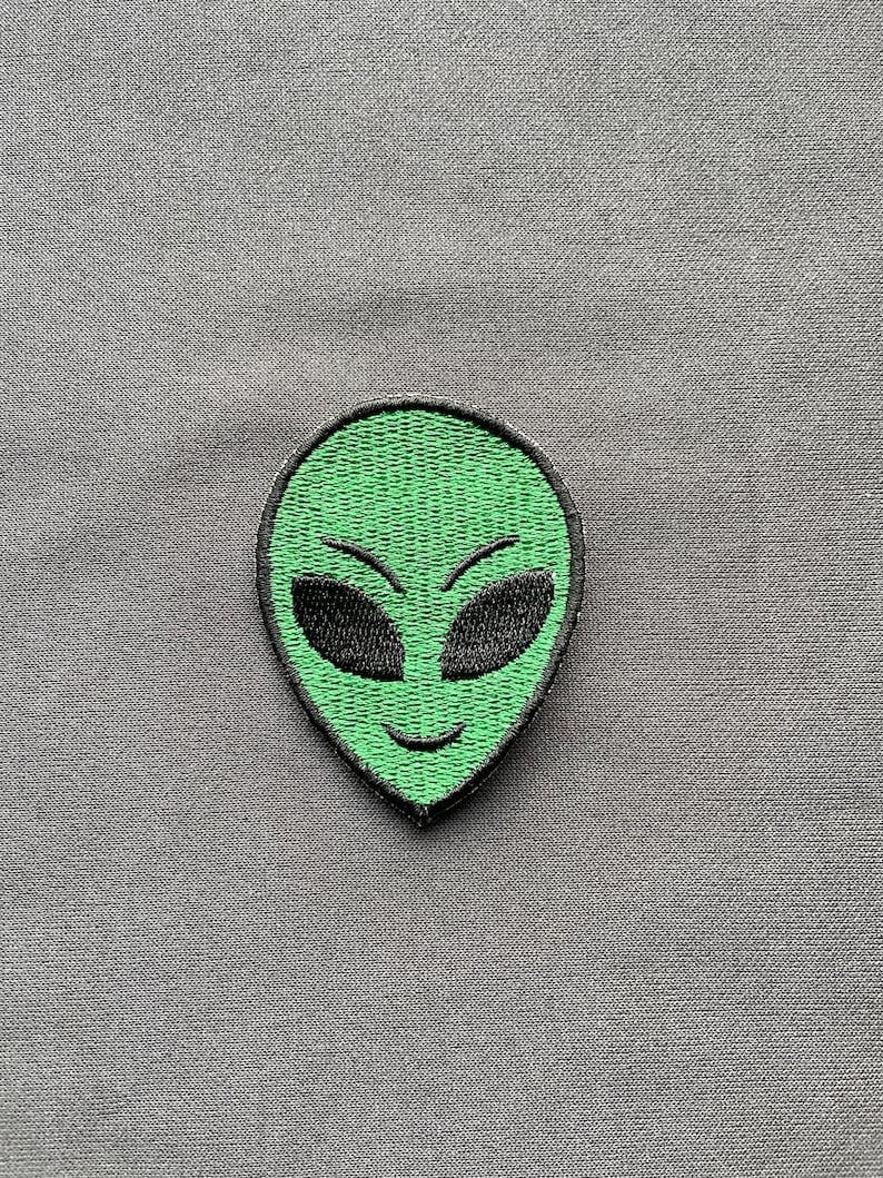 K&ouml;nnte beinhalten: Gestickter Aufn&auml;her mit einem gr&uuml;nen Alien-Gesicht mit schwarzen Augen und einem l&auml;chelnden Mund. Der Aufn&auml;her hat einen schwarzen Rand und befindet sich vor einem grauen Stoffhintergrund. Das Alien-Design ist ein beliebtes Thema.