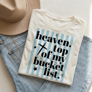 May include: Cream-colored t-shirt with the text "heaven. top of my bucket list." in black, set against a light blue striped background. The shirt is folded on top of blue jeans, with a tan hat and gold necklace visible.