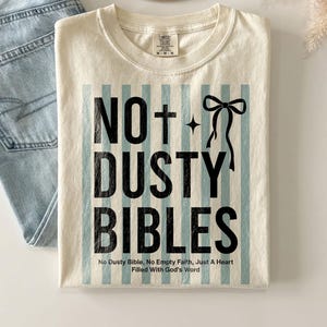 May include: Cream-colored t-shirt with the text "NO+ DUSTY BIBLES" in black, set against a backdrop of light blue vertical stripes. The shirt also features a cross and a bow graphic. The text "No Dusty Bible, No Empty Faith, Just A Heart Filled With God's Word" is printed at the bottom.