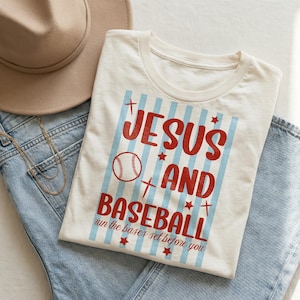 Pode incluir: Camiseta creme com as palavras "JESUS'S AND BASEBALL" em vermelho, com um gráfico de beisebol e listras azuis. O texto "run the bases set before you" está abaixo. A camisa está sobre jeans azuis.