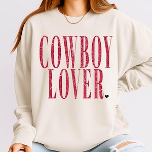 Puede incluir: Sudadera color crema con el texto "COWBOY LOVER" en letras rojas desgastadas. Un pequeño corazón negro está debajo del texto. La sudadera tiene cuello redondo y mangas largas.